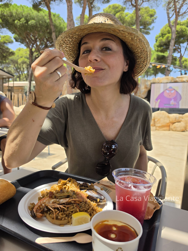 Una donna con cappello di paglia sorride mentre prende un boccone di riso, seduta a un tavolo all'aperto con un piatto di cibo e una bevanda.